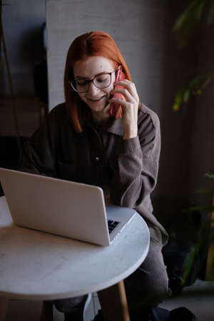 a red-haired freelance woman with glasses is talking on the phone, working remotely in a cafe. there is a laptop nearby.の写真素材