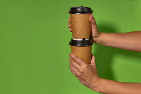 Close-up on a green background, womens hands hold two paper cups with coffee in a stand. Coffee or...