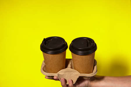 Close-up on a yellow background, womens hands hold two paper cups with coffee in a stand. Coffee or tea with you. Promotion two for the price of one, coffee delivery on a yellow backgroundの写真素材