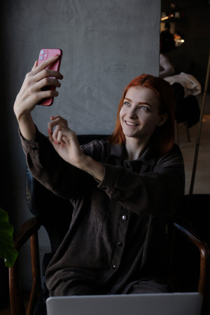 A red-haired woman takes pictures of herself on the phone while sitting in a cafe.の写真素材