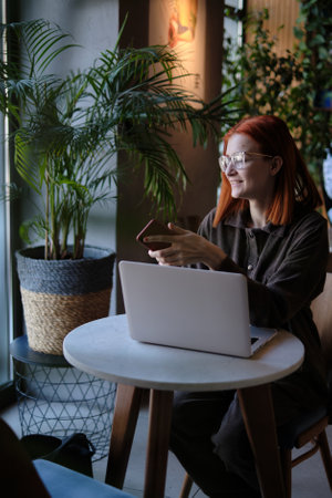 a red-haired freelance woman with glasses works remotely on a laptop in a cafe.の写真素材