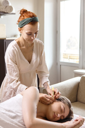 Beautiful woman ear receiving candle treatment. Therapist performing an ear wax treatment on a patient using candling. Naked woman receiving ear candle treatment from masseurの写真素材