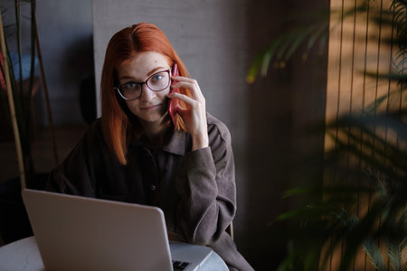 a red-haired freelance woman with glasses is talking on the phone, working remotely in a cafe. there is a laptop nearby.の写真素材