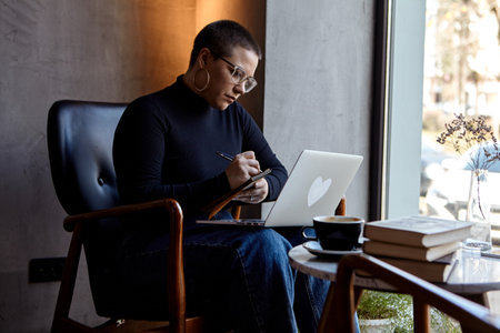 A young girl with glasses, with short hair is sitting in a cafe and working on a laptop. The girl looks at her laptop and makes notes in a notebook. The concept of distance learningの写真素材