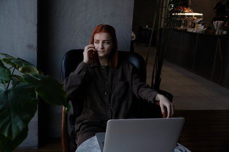 a red-haired freelance woman with glasses is talking on the phone, working remotely in a cafe. there is a laptop nearby.の写真素材