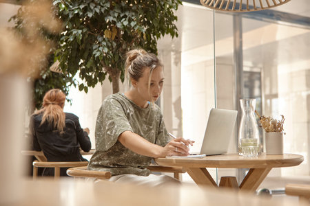 A young woman is sitting in a cafe and working on a laptop. The concept of freelancing and remote work or training. A woman makes notes in a notebook and looks at the laptop screenの写真素材