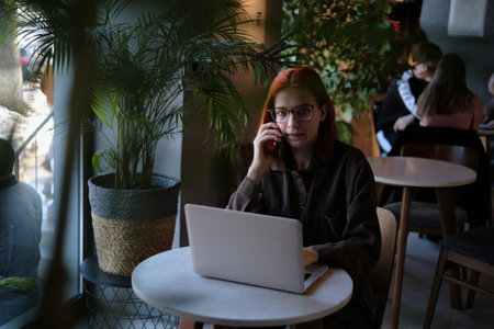 a red-haired freelance woman with glasses is talking on the phone, working remotely in a cafe. there is a laptop nearby.の写真素材
