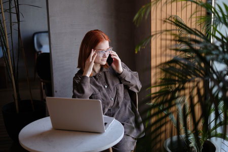 a red-haired freelance woman with glasses is talking on the phone, working remotely in a cafe. there is a laptop nearby.の写真素材