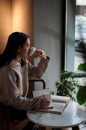 Shot of young asian female student sitting at table and writing on notebook. Young female student...