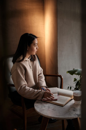 Shot of young asian female student sitting at table and writing on notebook. Young female student studying in cafe.の写真素材