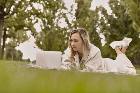 The woman is lying down using a laptop in the park. Smiling young student woman is using a laptop laying on grass.の写真素材