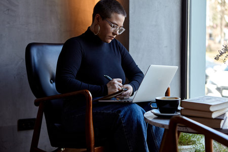 Young woman with short hair and nose piercing sits in a cafe. A woman is talking on the phone and work at a tablet. The concept of freelancing and remote work.の写真素材