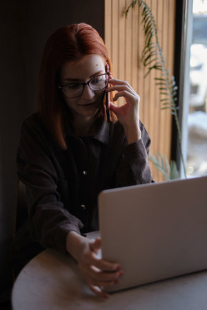 a red-haired freelance woman with glasses works remotely on a laptop in a cafe.xの写真素材