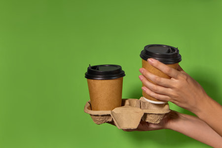 Close-up on a green background, womens hands hold two paper cups with coffee in a stand. Coffee or tea with you. Promotion two for the price of one, coffee delivery.の写真素材