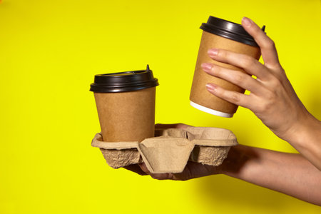 Close-up on a yellow background, womens hands hold two paper cups with coffee in a stand. Coffee or tea to go. Promotion two for the price of one, coffee delivery on a yellow backgroundの写真素材