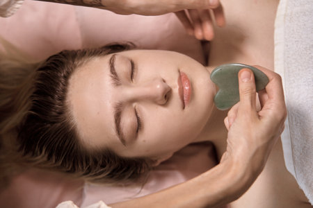 Young beautiful woman is getting a relaxing face Gua Sha Massage in wellness center. Masseur doing massage the head of an young beautiful girl woman in the spa salon close up macroの写真素材