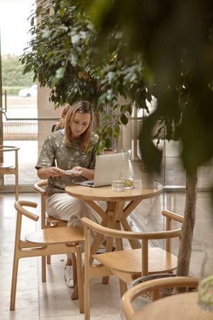 A young woman is sitting in a cafe and working on a laptop. The concept of freelancing and remote work or training. A woman makes notes in a notebook and looks at the laptop screenの写真素材
