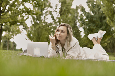 The woman is lying down using a laptop in the park. Smiling young student woman is using a laptop laying on grass.の写真素材