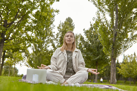 A woman sits cross-legged in the lotus position Sukhasana and meditates in the park. Attractive woman relieves stress by meditating in the parkの写真素材