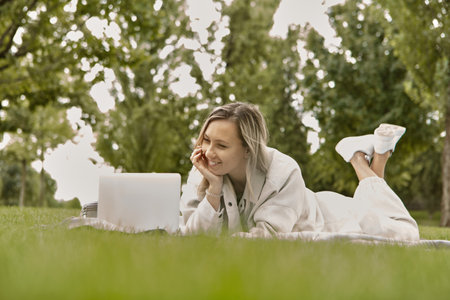 The woman is lying down using a laptop in the park. Smiling young student woman is using a laptop laying on grass.の写真素材