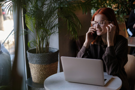a red-haired freelance woman with glasses is talking on the phone, working remotely in a cafe. there is a laptop nearby.の写真素材