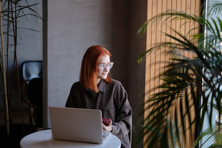 a red-haired freelance woman with glasses works remotely on a laptop in a cafe.xの写真素材