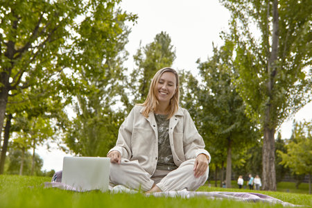 A woman is sitting on the grass in the park next to an open laptop. the concept of remote work or distance learning in an open spaceの写真素材