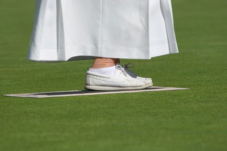 Ankles and feet of an elderly female wearing a long white skirt and white lawn bowling shoes, standing on a mat on a lawned area. の写真素材