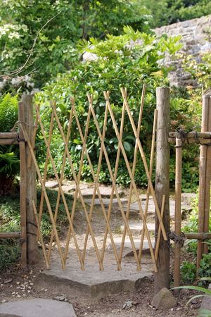 Bamboo gate of a diamond design, dividing a stone path in a garden with shrubs to the rear.の写真素材