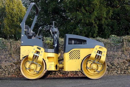 Steam roller standing idle on a path with an old stone wall and trees to the rear.