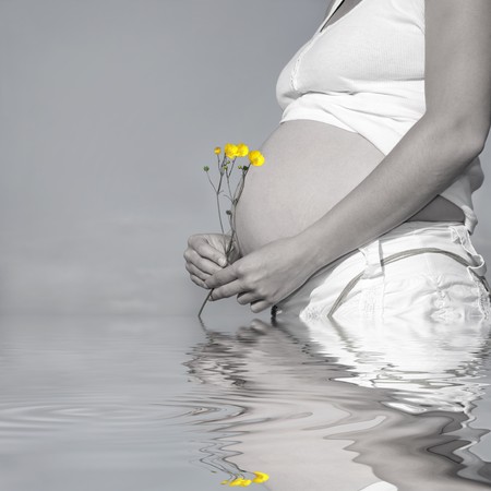 Pregnant woman holding a bunch of buttercups with exposed bare stomach. Desaturated with only the flowers saturated and reflection over rippled water.の写真素材