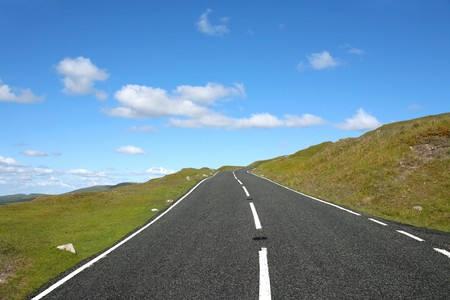 Uphill rural road with grass verges on either side and a blue sky with clouds. 
