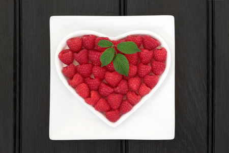 Raspberry super food fruit in a heart shaped bowl on a white square dish over wooden black background.の写真素材
