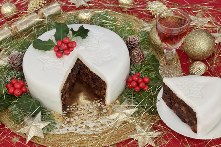 Iced christmas cake and slice with glass of sherry, holly, ivy, fir and gold bauble decorations on red background with tinsel strands.の写真素材