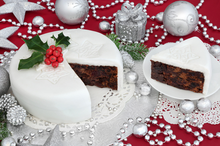 Traditional iced christmas cake and slice with holly, bauble decorations and silver foil wrapped chocolates on red background.の写真素材