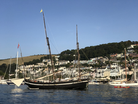 Dartmouth, Devon UK 28th August 2017. Historical old Grayhound lugger sailing boat moored on the River Dart with the quaint hillside town of Darthaven to the rear. Used for education and sailing experiences.のeditorial素材