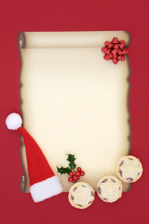 Letter to Santa Claus with blank parchment paper, santa hat, mince pies, bow and holly berry leaf sprig on red background. Christmas Eve concept.の写真素材