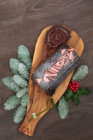 Chocolate yule log cake with traditional winter flora of snow covered fir, holly and mistletoe on rustic wooden board and oak table background. Sweet food for Christmas.の写真素材