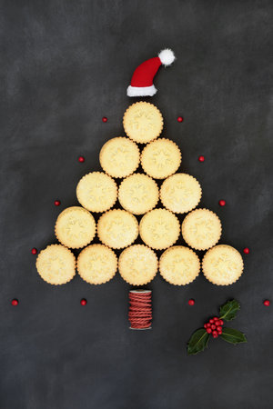 Abstract Christmas tree concept shape with mince pies, santa hat, winter holly with red berries on grunge gray background. Surreal food design for the Xmas and New Year holiday season.の写真素材