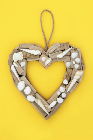 Driftwood heart shaped wreath with seashells. Natural rustic beautiful holiday and travel destinations object. Symbol for Mothers Day, anniversary, birthday, Valentines Day. On yellow background.の写真素材