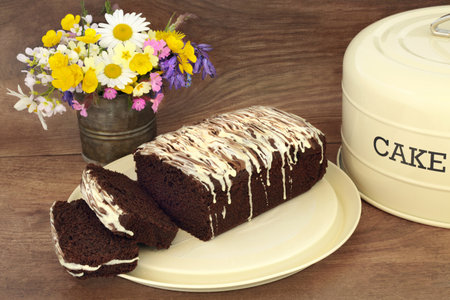 Chocolate chip cake with icing drizzle on metal tray with lid on rustic wood background with Spring floral wildflower arrangement. Homemade sweet food  for afternoon tea.の写真素材