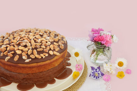 Peanut cake decorated with peanuts and gooey caramel sauce on pink background with summer flowers. Homemade afternoon tea composition.の写真素材