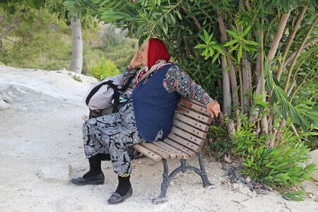 PAMUKKALE, TURKEY - AUGUST 22, 2017: An elder woman seats on a bench, relaxing and enjoying a beautiful day. A popular way, among people who live in Pamukkale, is to spend their free time, outdoors.のeditorial素材
