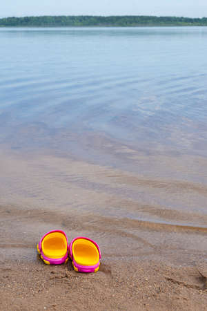 Children's pink clogs stand on a sandy beach near the water.の写真素材