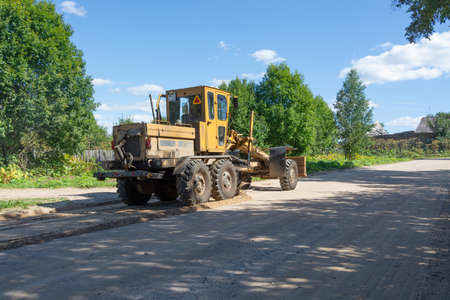 The grader levels a dirt road in a rural area with a special blade with a knife, which is mounted on...