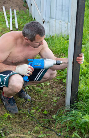 At the dacha, a man sets up a fence, drills a metal pole with a drill.のeditorial素材