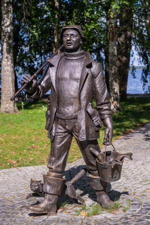 Ostashkov, Tver region, Russia - 08/15/2023. Monument to the fisherman in Ostashkov. A bronze figure of a fisherman with a fishing rod, a bucket of fish and a cat, installed in theの写真素材