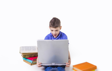 Portrait of cute teen schoolboy sitting near books and typing on laptop keyboard. Happy and surprised little boy, caucasian model isolated on white studio background. Education, study, studying, learning conceptの写真素材