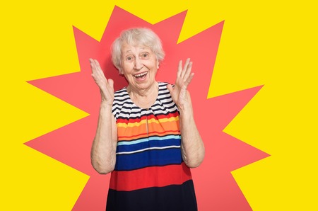 Old smiling woman with surprised expression on her face on red studio background. Human emotions concept. Positive emotional old lady standing indoorの写真素材