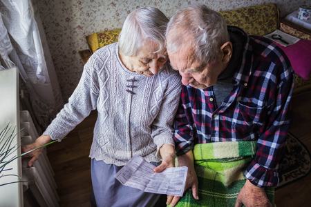 The senior woman and man holding gas bill in front of heating radiator. Payment for heating in winter.の写真素材